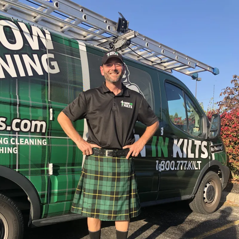Man in a green plaid kilt and black shirt stands smiling in front of a Men In Kilts service van with ladder on top.