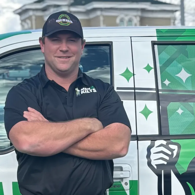 Man in black Men In Kilts shirt and cap stands with arms crossed beside a branded white and green service van.
