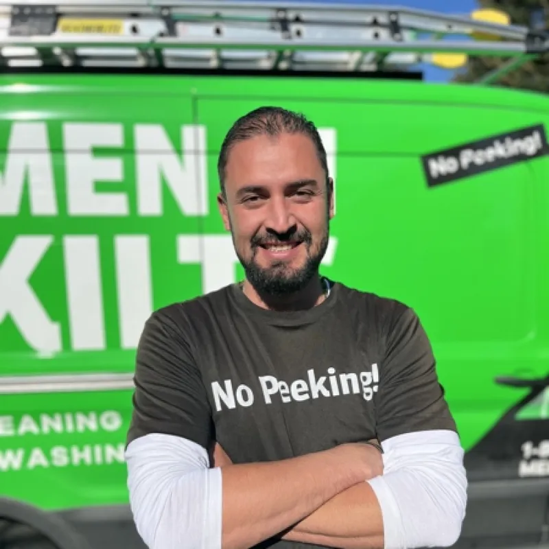 Smiling man with crossed arms in No Peeking! shirt standing in front of a green maintenance service van.