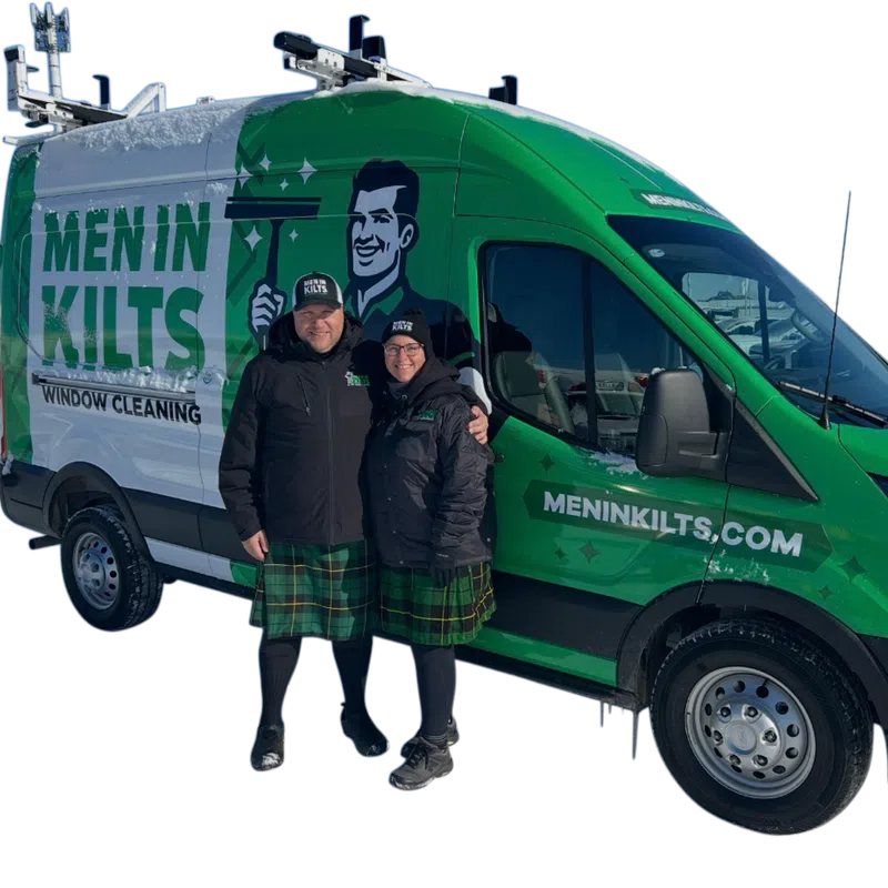 Two window cleaners wearing kilts standing next to a green Men in Kilts window cleaning van in snow.