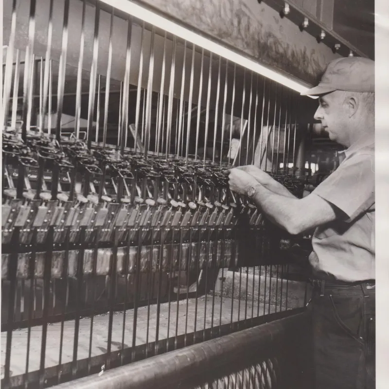 Worker operating and inspecting textile machinery with numerous spools and threads in a factory setting.