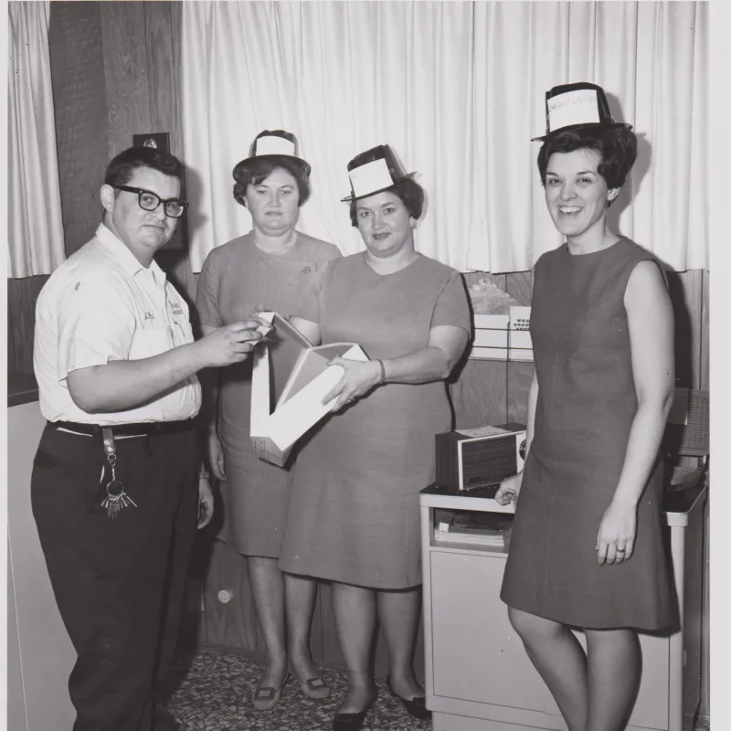 Four people in 1960s attire with paper hats, holding a box, in an office with wood paneling and tiled floor.