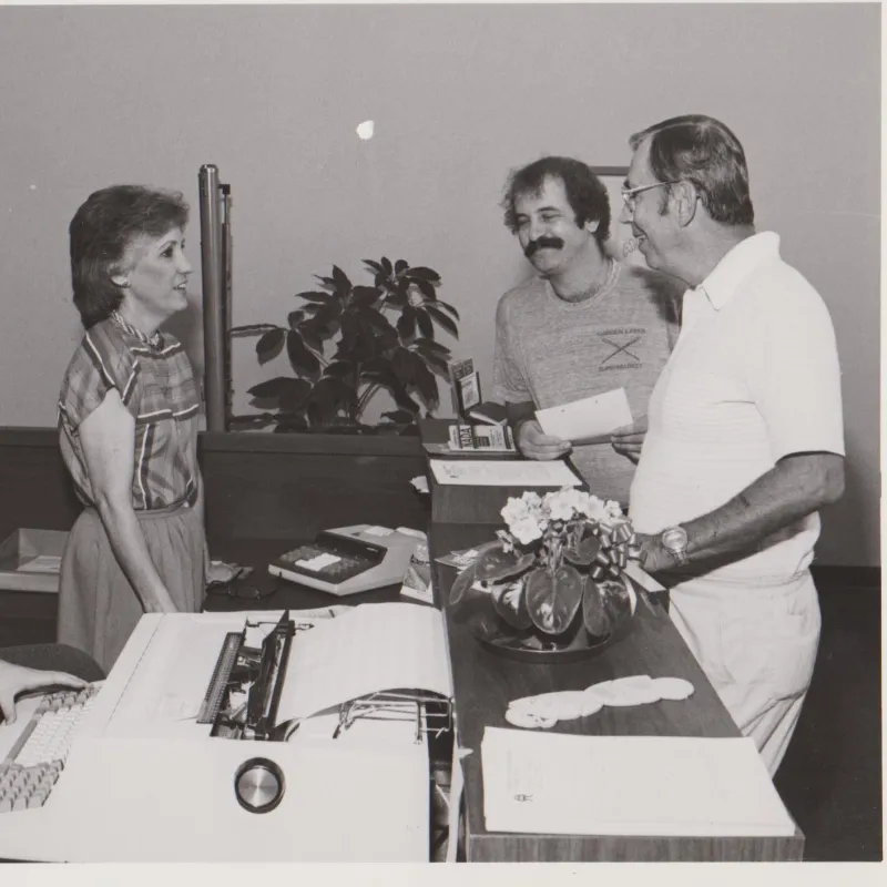 Vintage office scene with four people around desks, including typewriter, calculator, and paperwork in black and white photo.