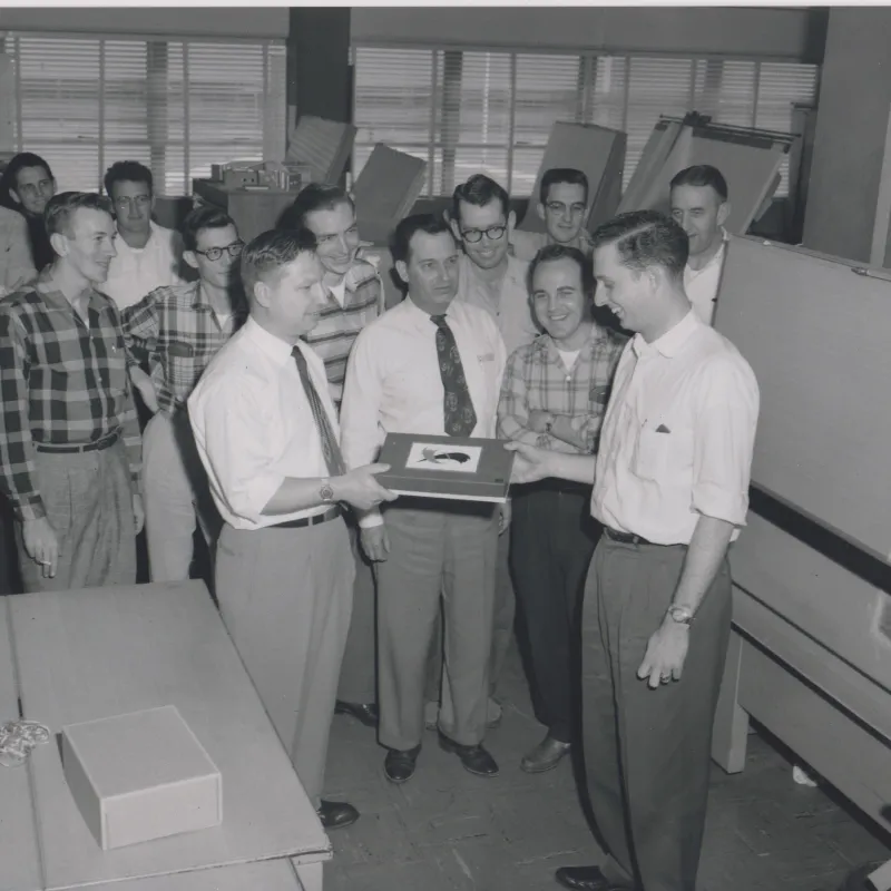 Group of men in vintage office setting presenting a framed item, smiling and engaged in discussion.