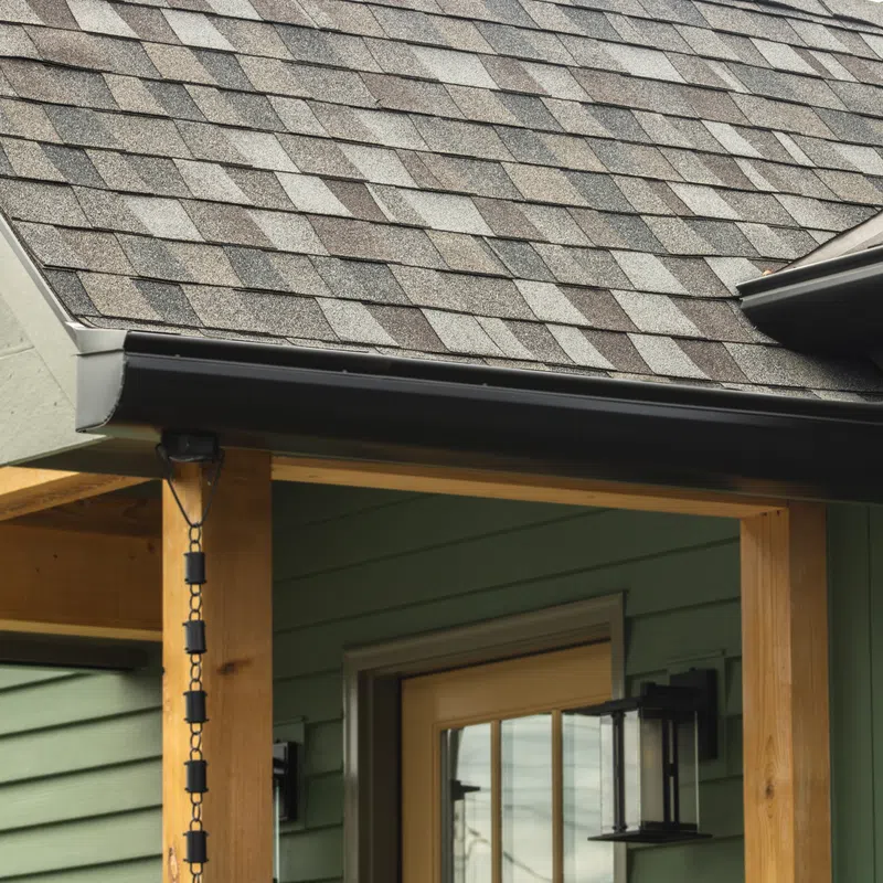 Close-up of house roof with gray shingles, wooden porch frame, green siding, and black rain chain by the gutter.