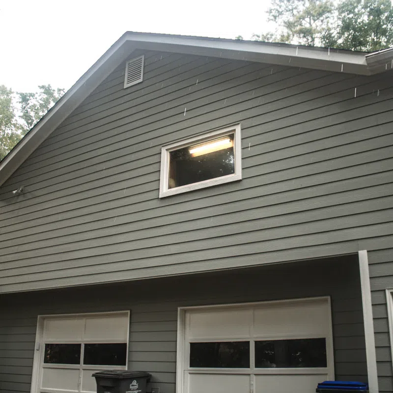 Gray two-car garage exterior with white door frames and a lit window on upper floor, surrounded by trees.