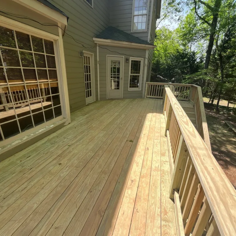 Spacious wooden deck attached to a house with railing and large windows, surrounded by trees and sunlight.