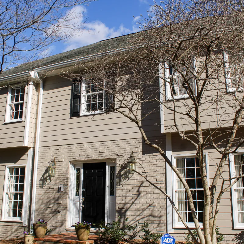 Two-story beige house with black front door, white-framed windows, leafless trees, and clear blue sky on a sunny day