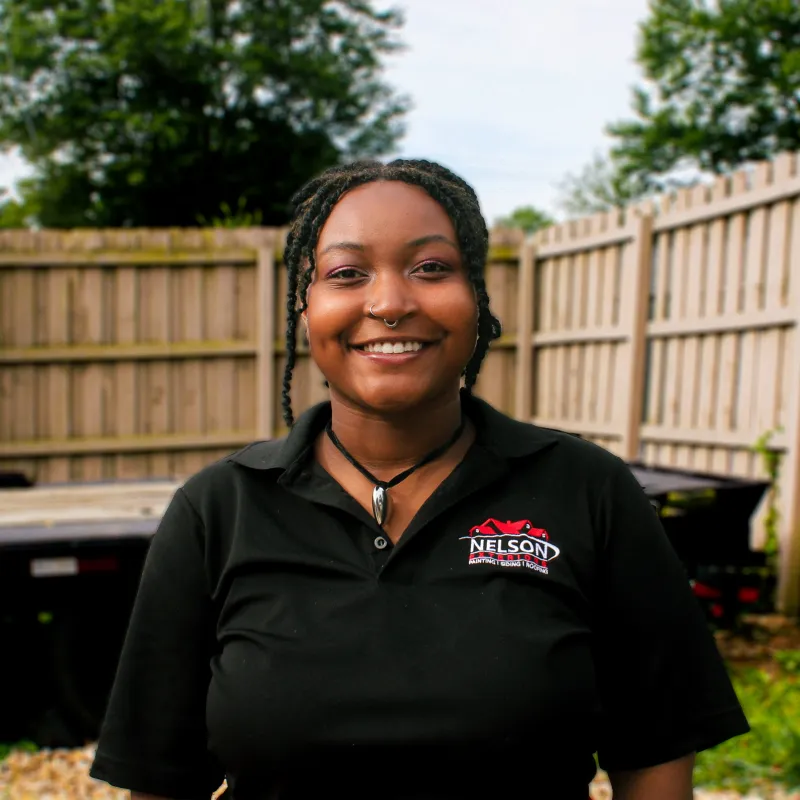 Smiling woman with braided hair wearing a black Nelson Rental Services polo shirt outdoors by a wooden fence