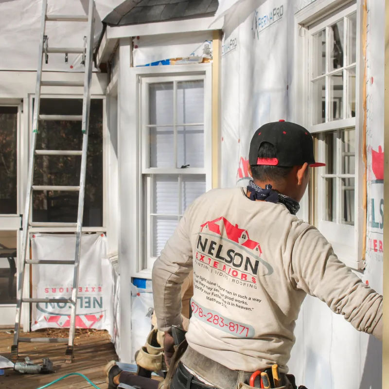 Worker from Nelson Exteriors installing siding on a home exterior under renovation with ladders nearby.