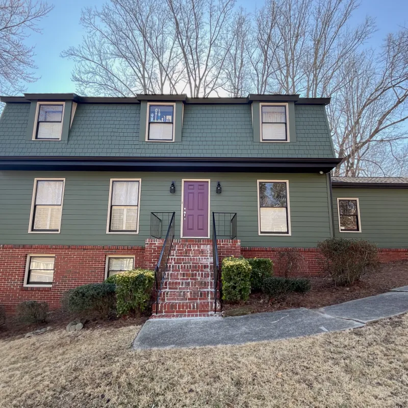 Green and brick two-story house with purple front door, staircase, shrubs, and leafless trees under blue sky.