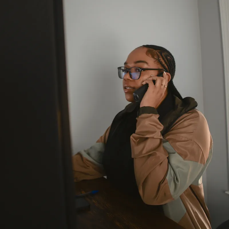Person with braided hair and glasses talking on phone at desk in a casual indoor setting.
