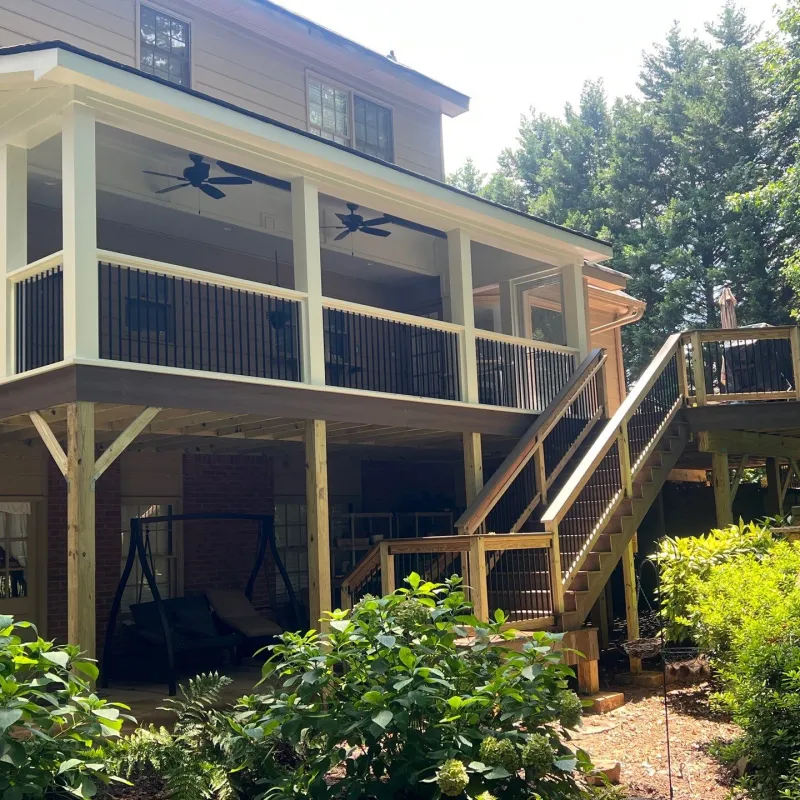 Two-level wooden deck with screened upper porch and outdoor stairs surrounded by green shrubs and trees.
