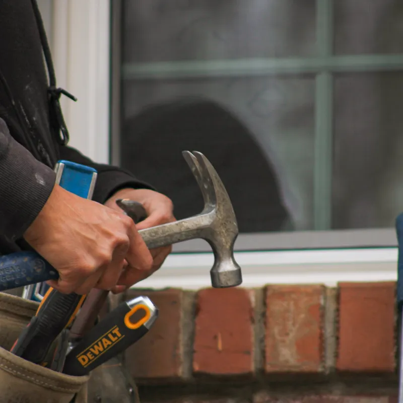 Construction worker holding a hammer with tools in a belt near a brick wall and window.