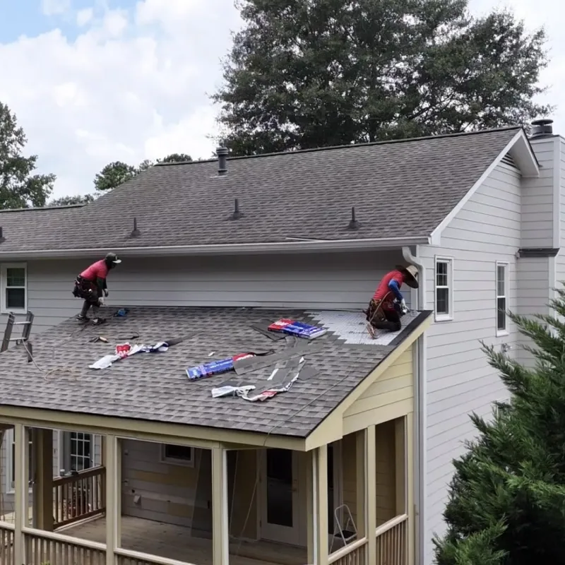 Two workers installing new shingles on a house roof on a sunny day surrounded by trees in a suburban neighborhood.