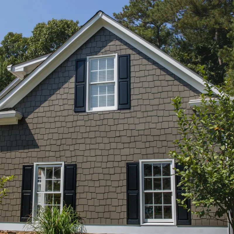 Gray shingle siding house with white windows and black shutters surrounded by trees under a clear sky