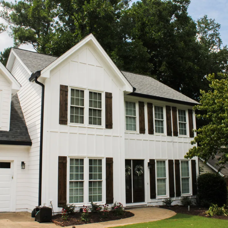 Two-story white house with dark shutters, gabled roof, attached garage, and surrounding greenery under a blue sky.