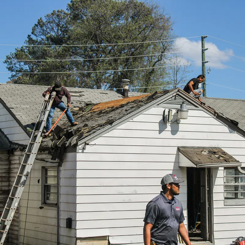 Workers removing damaged roof shingles from a white house under clear blue sky during roof repair work.