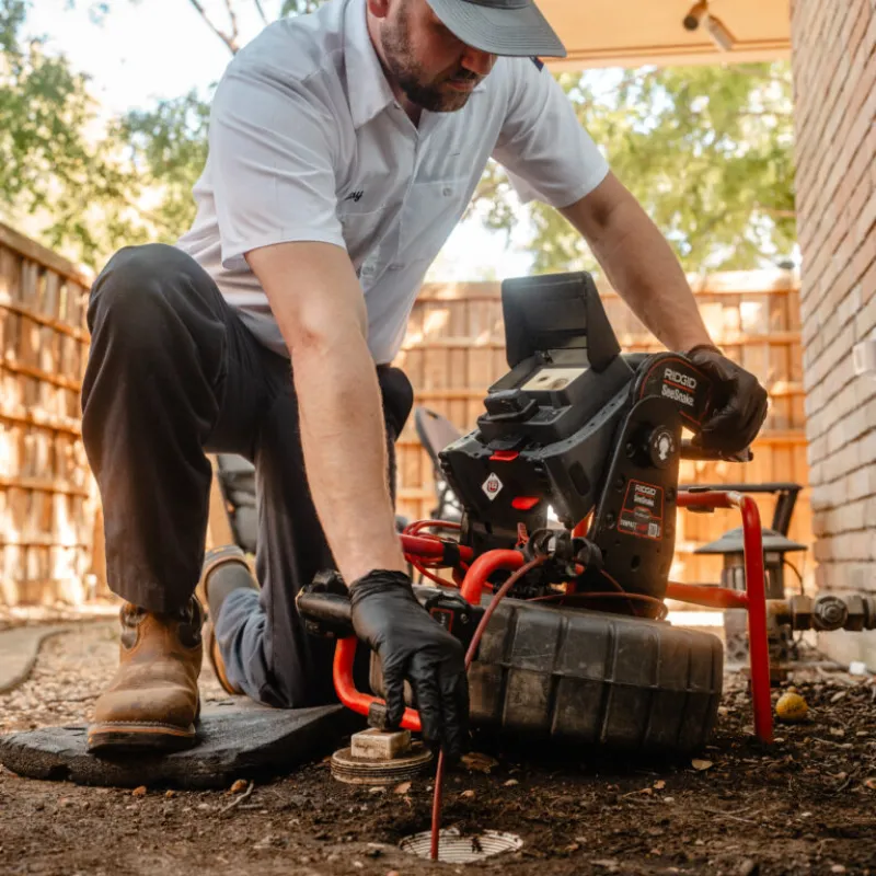 Plumber in uniform using a drain inspection camera outdoors near a house while kneeling on the ground.