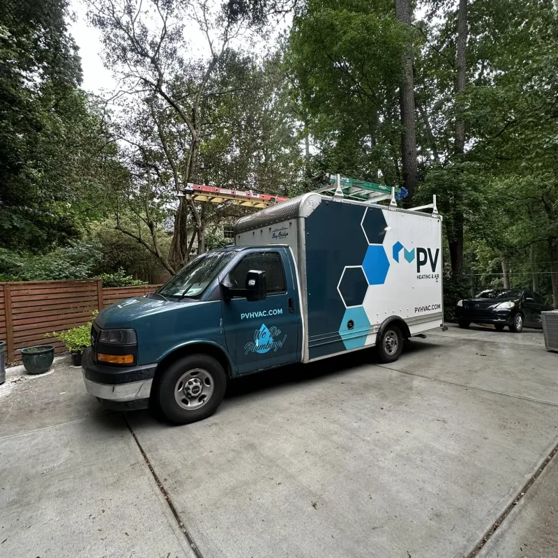 Blue and white PV HVAC plumbing service van parked on a concrete driveway in a green wooded residential area.