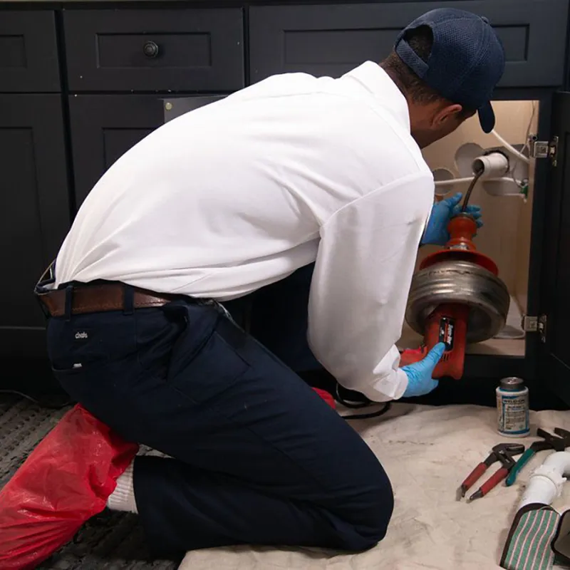 Plumber in white shirt and gloves kneeling while fixing a kitchen sink drain with tools nearby