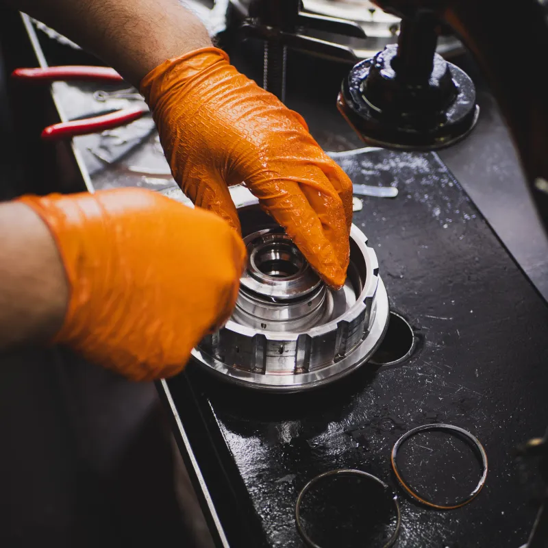 Technician wearing orange gloves assembling or repairing a mechanical metal component on a workbench.