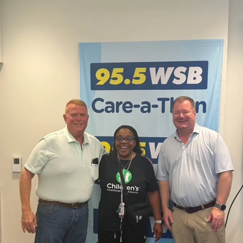 Three people smiling and posing in front of a 95.5 WSB Care-a-Thon banner in a radio station studio.