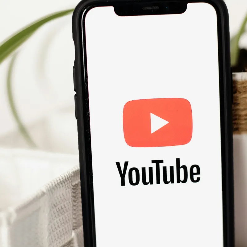 Smartphone displaying YouTube logo with red play button on white screen, next to a plant and white textile.