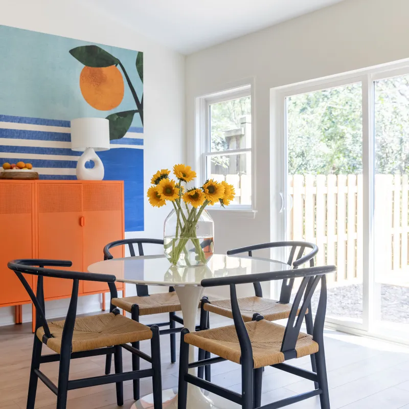 Bright dining room with glass table, black chairs, orange cabinet, sunflower centerpiece, and large windows.