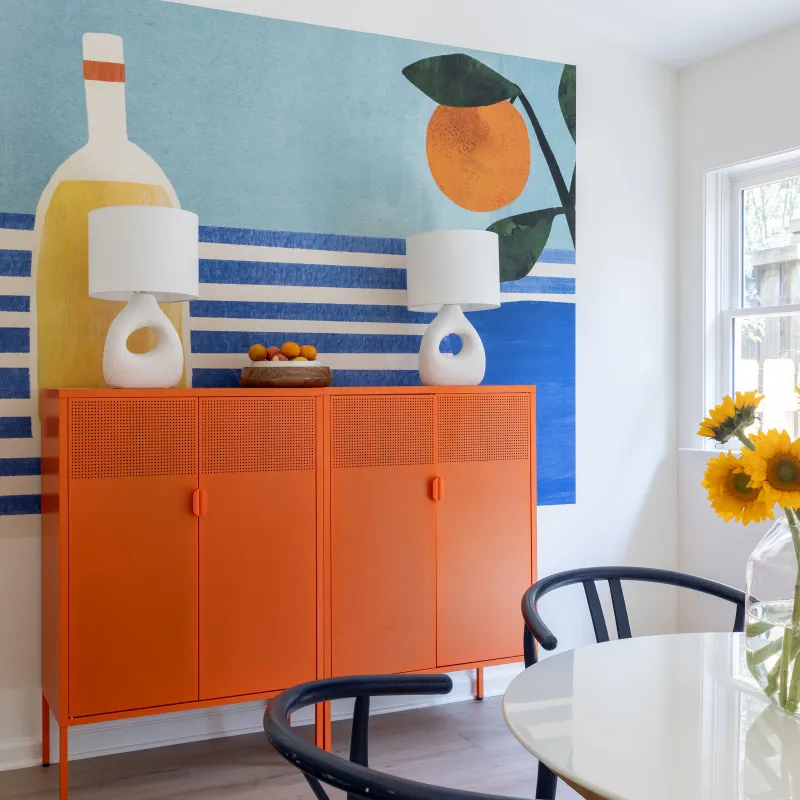 Bright dining room with orange sideboard, large colorful wall art, and sunflowers in glass vase on white table.