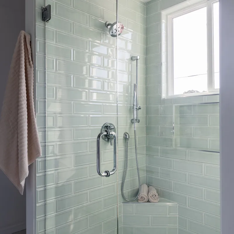 Modern bathroom shower with glass door, light green subway tiles, chrome fixtures, and rolled towels on bench.