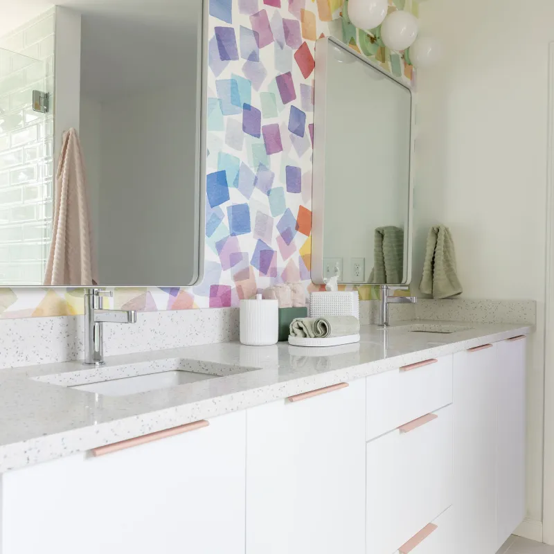 Modern bathroom with double sink, speckled white countertop, colorful geometric wall tiles, and round mirrors.