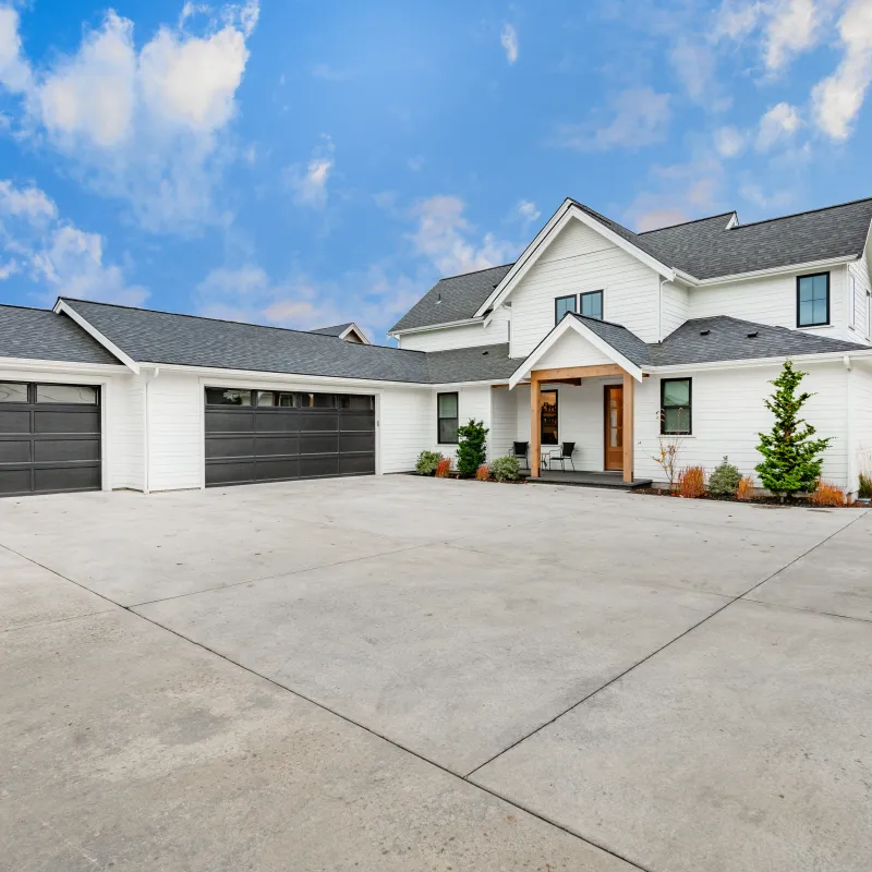 Modern white two-story house with black garage doors and expansive concrete driveway under blue sky.