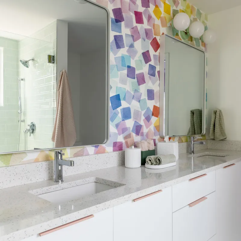 Bright modern bathroom with colorful geometric wall art, dual sinks, large mirrors, and white cabinetry.