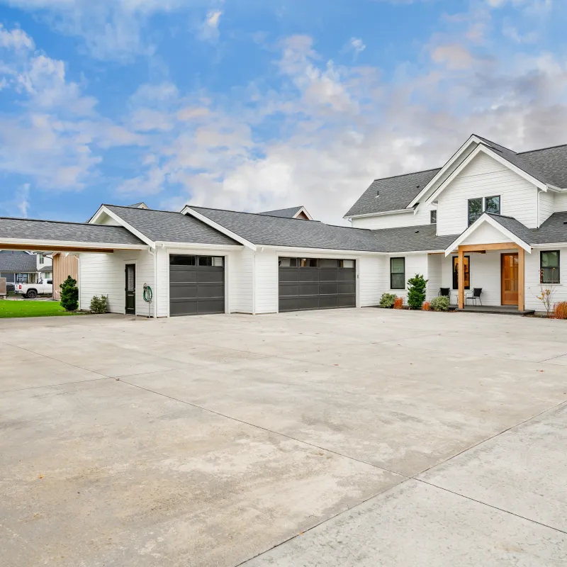 Modern white two-story house with multiple gray garages and large concrete driveway under a partly cloudy sky.