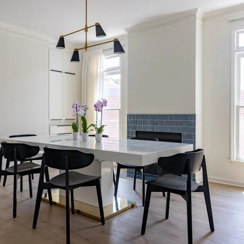 Modern dining room with white table, black chairs, hardwood floor, large windows, and a sleek blue tiled fireplace.