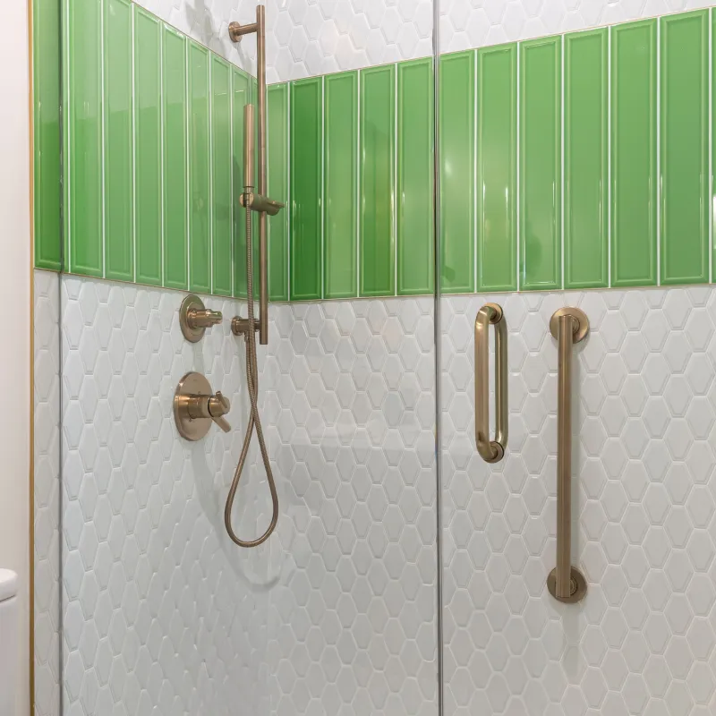 Modern shower with white hexagon tiles, green accent tiles, brass fixtures, and glass door with handle.