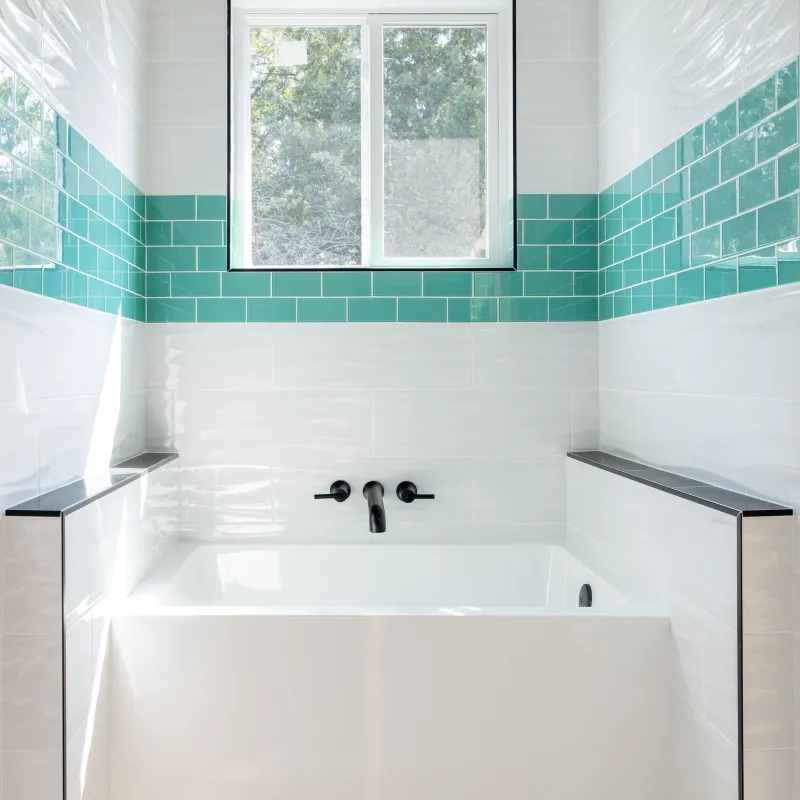 Modern white bathtub with black fixtures under window, surrounded by white and teal subway tiles in bathroom.
