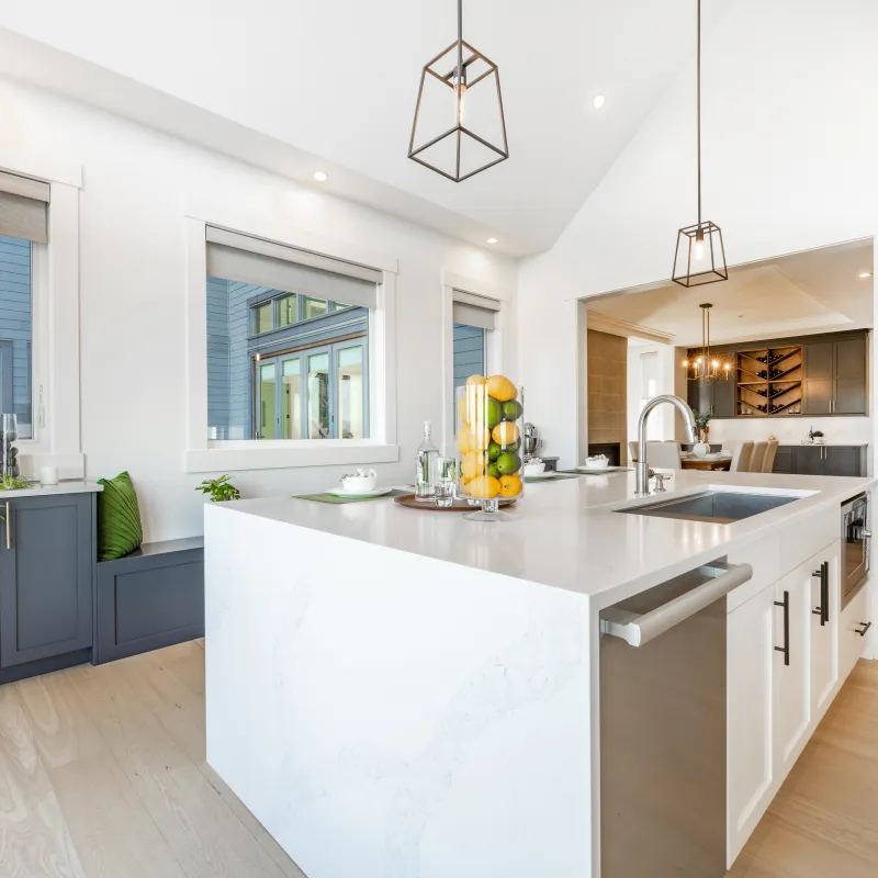 Bright modern kitchen with marble island, pendant lights, gray cabinets, wooden floors, and large windows.