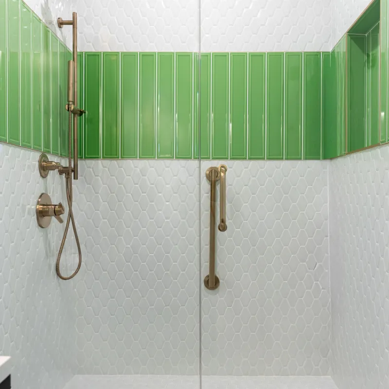 Modern shower with green rectangular tiles, white hexagonal tiles, brass fixtures, and glass door.