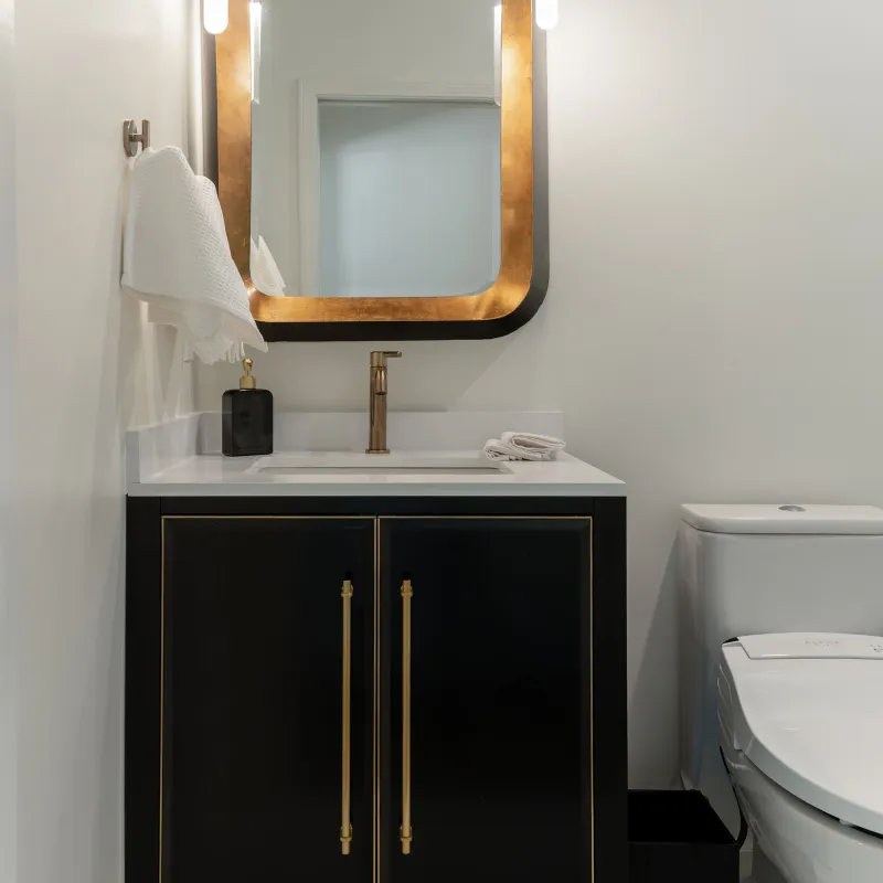 Modern bathroom vanity with black cabinet, gold-framed mirror, wall lights, and white countertop sink