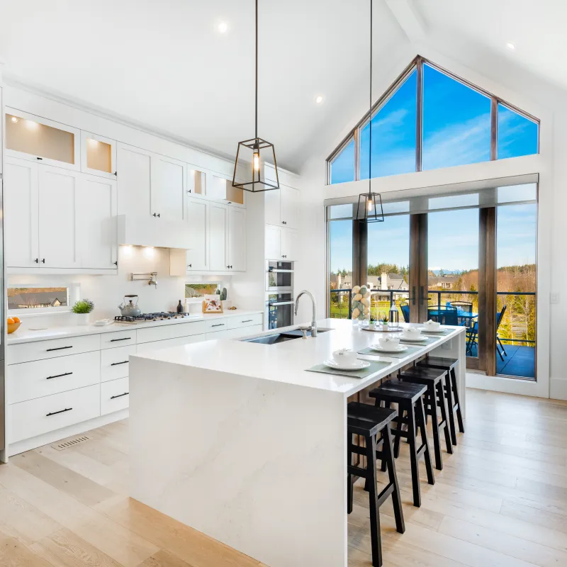Bright modern kitchen with white cabinets, marble island, black stools, and large windows overlooking outdoor balcony.