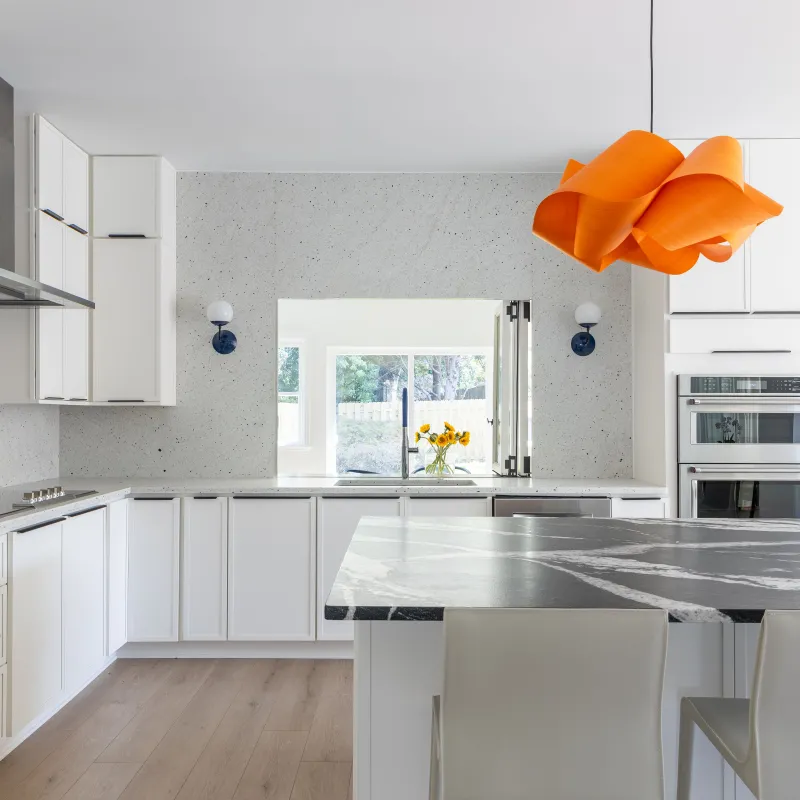 Modern white kitchen with marble island, stainless steel appliances, wooden floor, and orange pendant light.