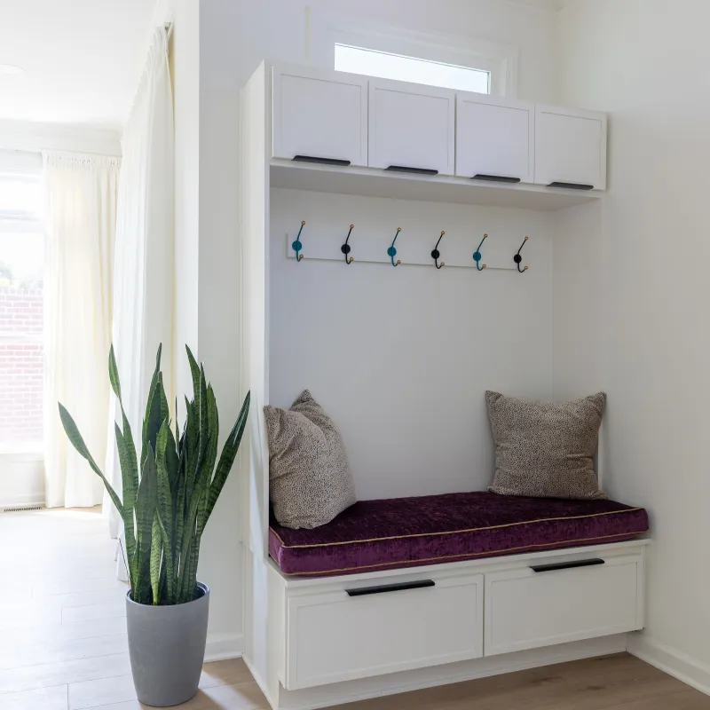 Bright entryway with white built-in bench, hook rack, cushions, and potted plant next to a white door and dining area.