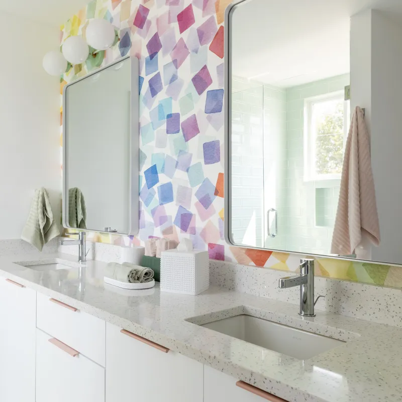 Bright modern bathroom with white double vanity, speckled countertop, colorful geometric tile backsplash, and rectangular mirrors.