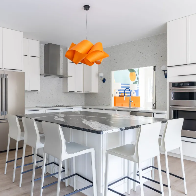 Modern white kitchen with large marble island, six white chairs, stainless steel appliances, and orange pendant light.