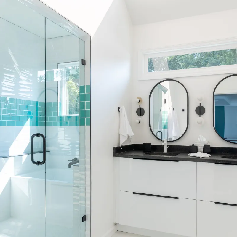 Bright modern bathroom with glass shower, turquoise tiles, black countertop, dual oval mirrors, and skylight