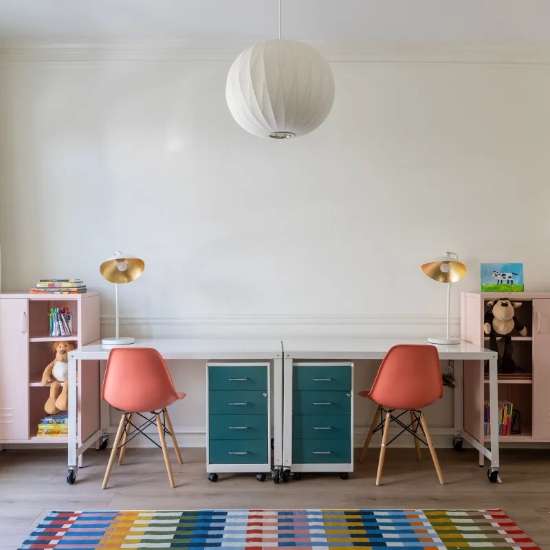 Minimalist twin study desks with coral chairs, teal drawers, pink lockers, colorful rug, and large window in bright room