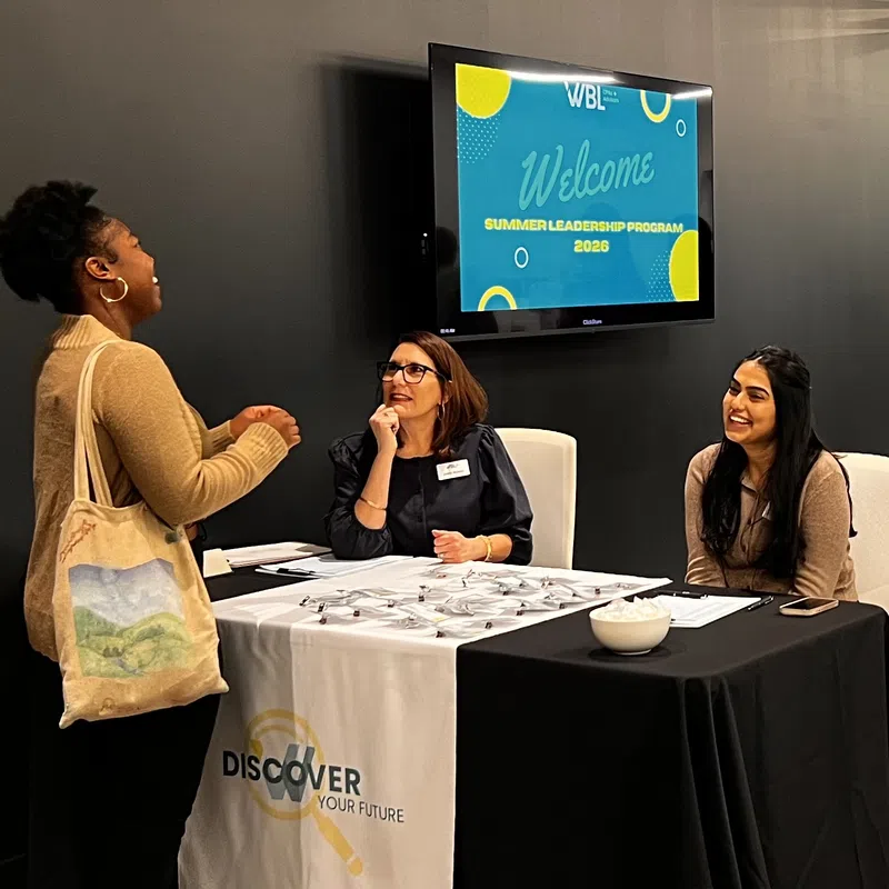 Three women engaged in conversation at a Summer Leadership Program 2026 registration table with welcoming signage.