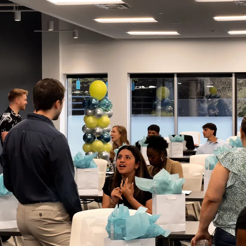 People socializing in a decorated event space with gift bags and balloons in a modern office setting.